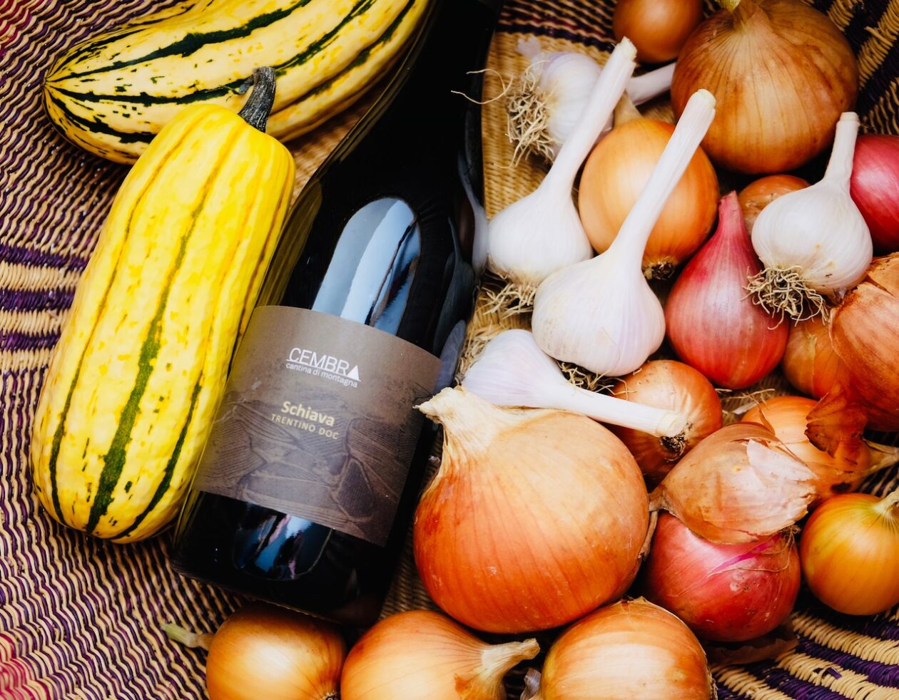 A bottle of Schiava wine lying in a basket with squash, onions, and garlic.