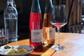Two bottles of Ryme Cellars wines on a table, with wine in a glass and a small cheese plate.