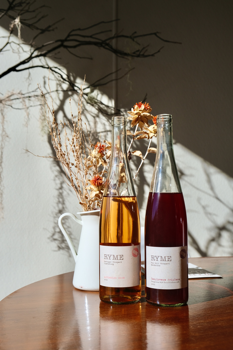 Two bottles of wine from Ryme Cellars, on a table with a vase of dried flowers.
